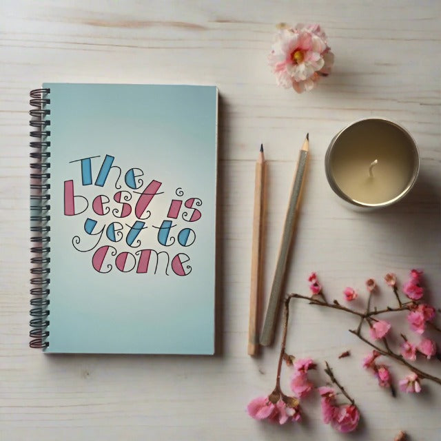 Notebook with writing on cover that reads - the best is yet to come. Pencils, candle and blossom next to the notebook