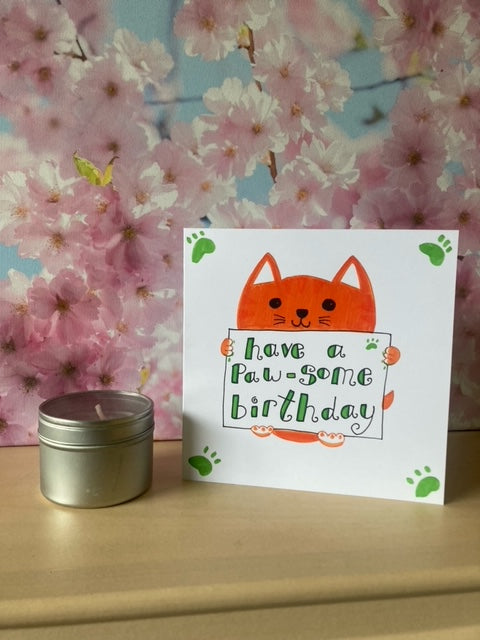 Handmade card with ginger cat holding a sign that says have a paw-some birthday. Sat on shelf with candle and blossom tree picture in background